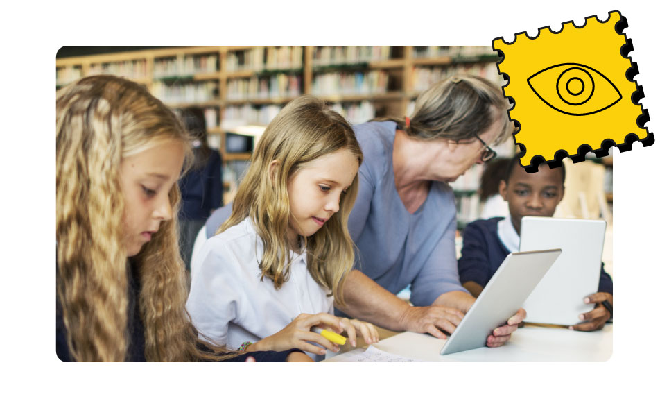 A researcher holding a tablet speaks to children in a school library.