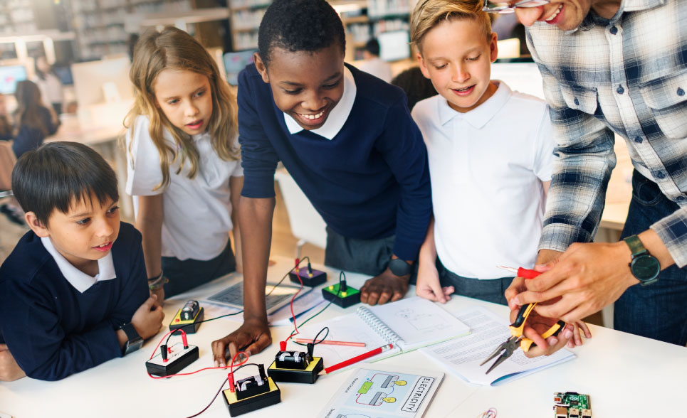 Four school children intently watching a teacher explaining electricity using circuit boards.