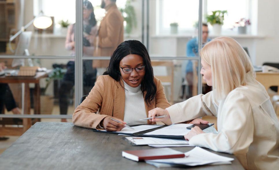 Two women discussing paper work at a large table. More people cann be seen working behind them in another part of the office.