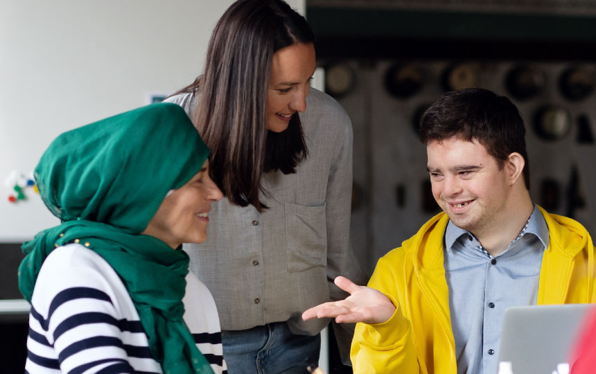 Two women, one wearing a headscarf and a man with Down-syndrome discuss ideas together in a community space.