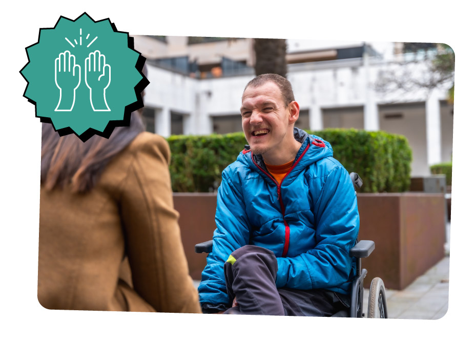 A person in a blue jacket sitting in a wheelchair is smiling and engaging in conversation with another person wearing a brown coat in an outdoor urban setting.