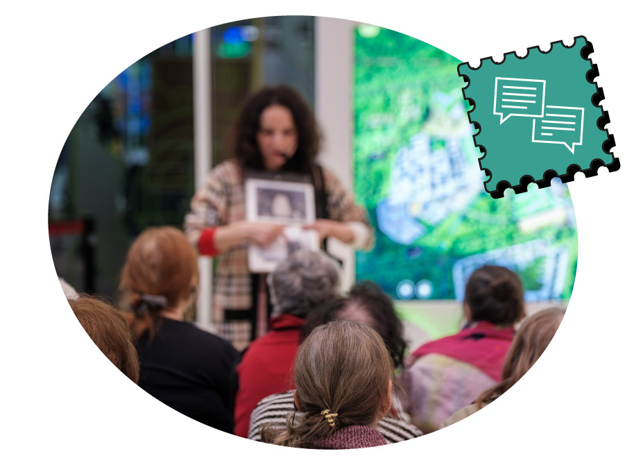 A person with curly hair and a patterned sweater is holding a photo and speaking to an audience of seated people in a room with a large screen displaying a map.