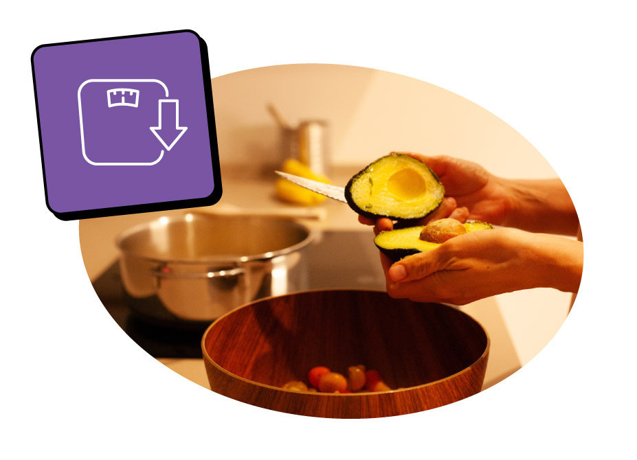 A person is holding a halved avocado over a wooden bowl with grapes inside in a kitchen setting with a pot on the stove.