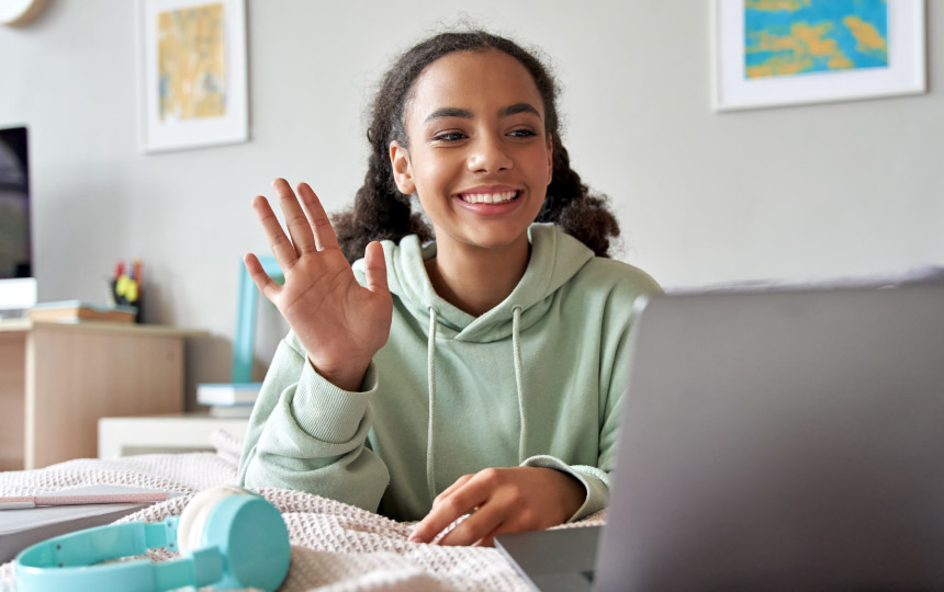 A young girl using a laptop, smiling and waving to others on a group call