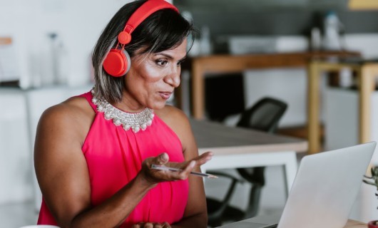 A transgender woman wearing red headphones in front of her laptop, taking part in a video meeting.