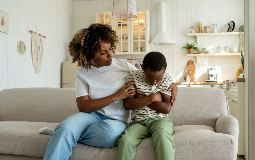 A mother sits beside her son who has his arms crossed and head down in an unhappy manner.