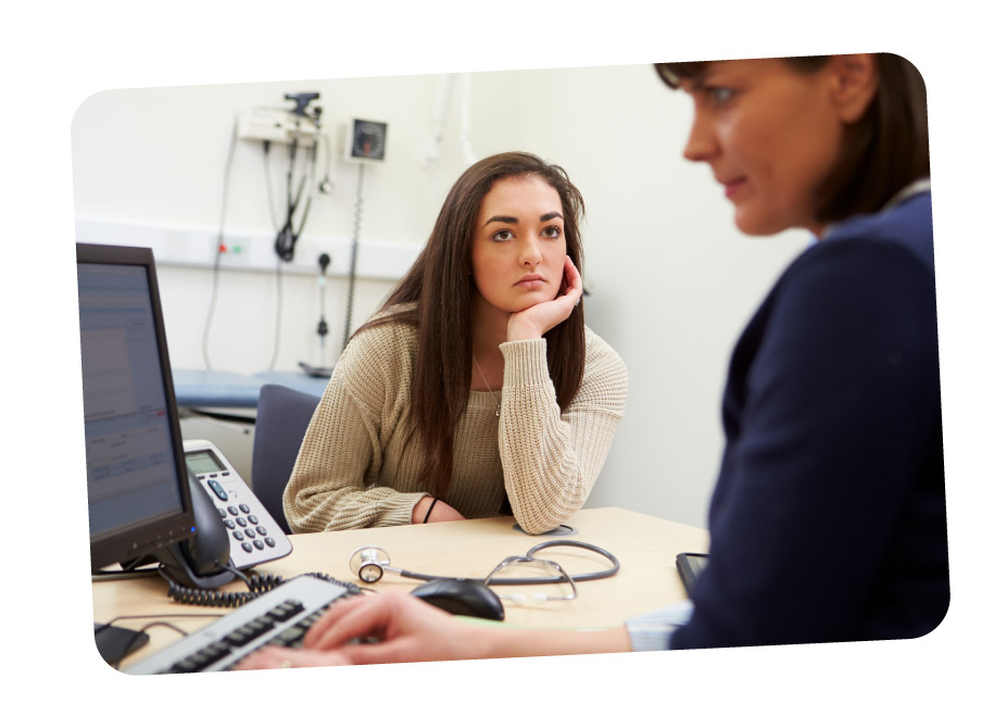 A woman with long hair wearing a beige sweater sits at a desk looking attentively at another person who is typing on a keyboard in a medical setting.