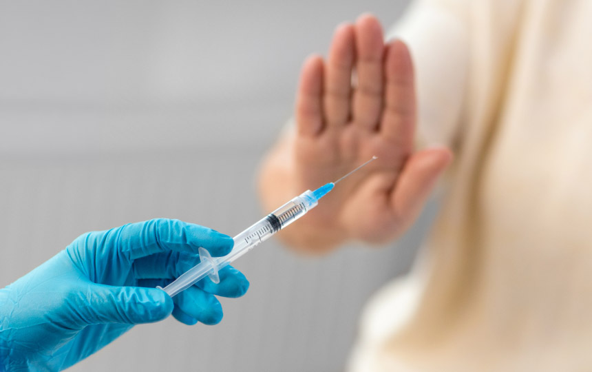 A person wearing blue gloves holds a syringe while another person raises their hand in a gesture of refusal.