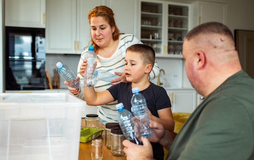 Three people in a kitchen are sorting and holding empty plastic bottles near a large clear storage bin on a wooden table.