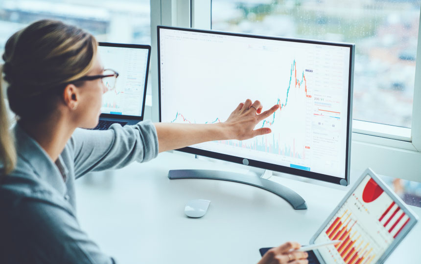 A woman in a brightly lit room points to graphs and statistics on her computer monitor