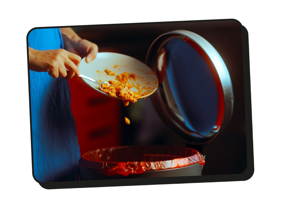 A woman wearing a blue dress scraping baked beans off a plate into a open bin.
