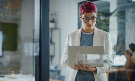 Person with short, pink hair in white blazer. Standing and looking at laptop. Set in a contemporary business environment.