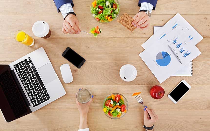 Two people are sat eating salads, with laptops, phones and paperwork on the table.
