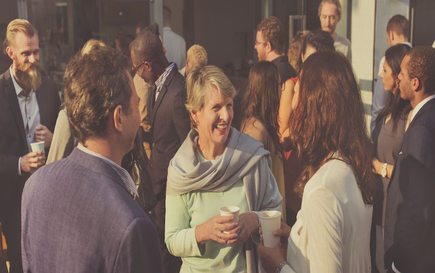 A small crowd of people are stood in various groups, talking to each other whilst having a drink.