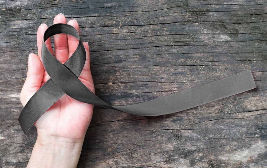 A hand resting on a wooden table holds a black, skin cancer awareness ribbon.