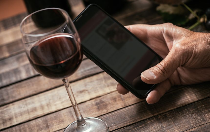 A hand holds a smart phone at a wooden table, with a small glass of red wine next to it.