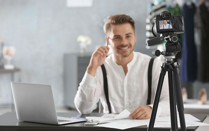 A man sat at a desk filming himself on a camera on a tripod, with his laptop in front of him.
