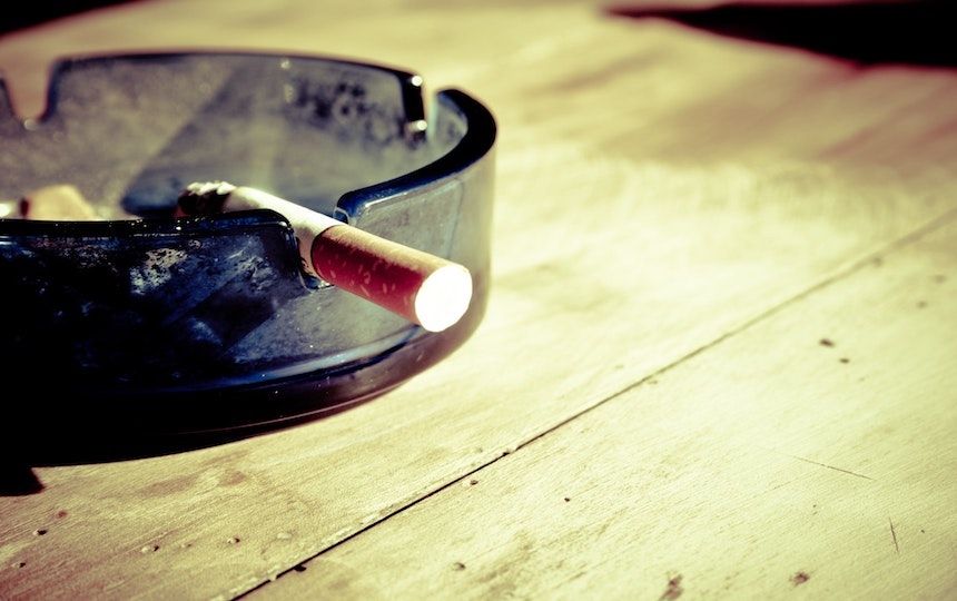 An ashtray is on a wooden table, with a half smoked cigarette hanging out of the ashtray.