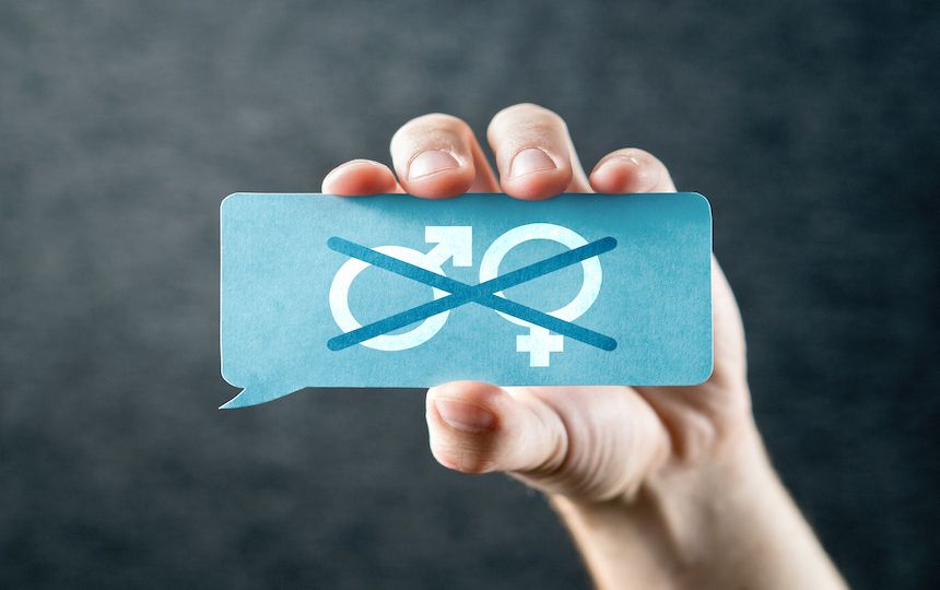 A hand holding a blue speech bubble card, with a female and male symbol, which has been crossed out.
