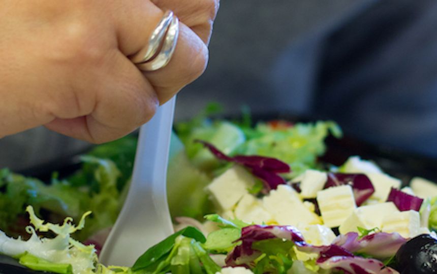 A hand is holding a plastic fork in a plastic container which is full of fresh salad.