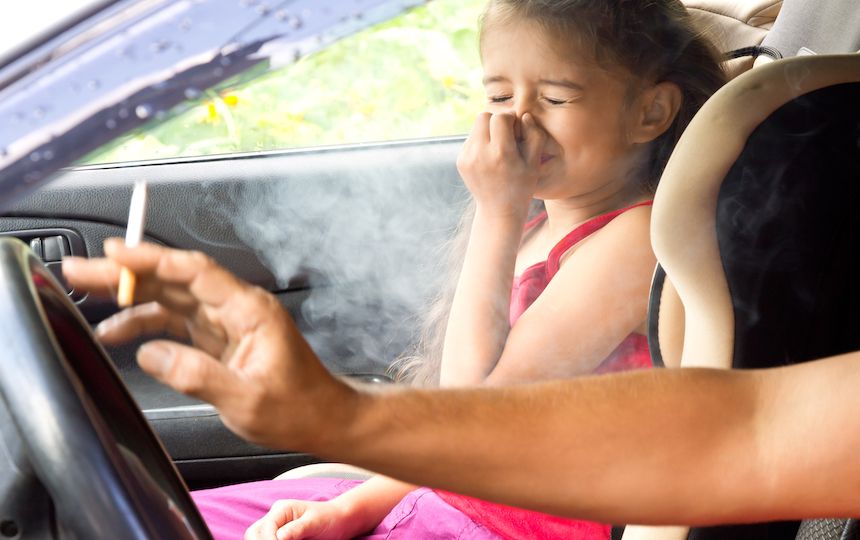 A child is sat in a car's passenger seat holding her nose against the driver's cigarette smoke.