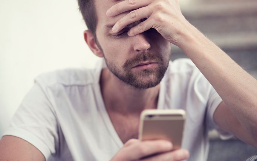 A man is looking at his smartphone whilst holding his head in his hand.