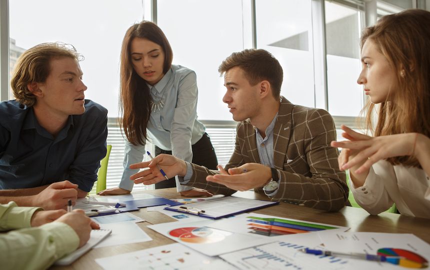 Five smartly dressed people are having a discussion around a table topped with charts and documents.