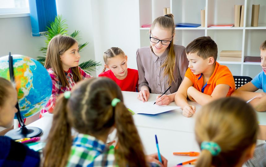A woman sits at a table in a classroom setting, surrounded by children watching her draw.