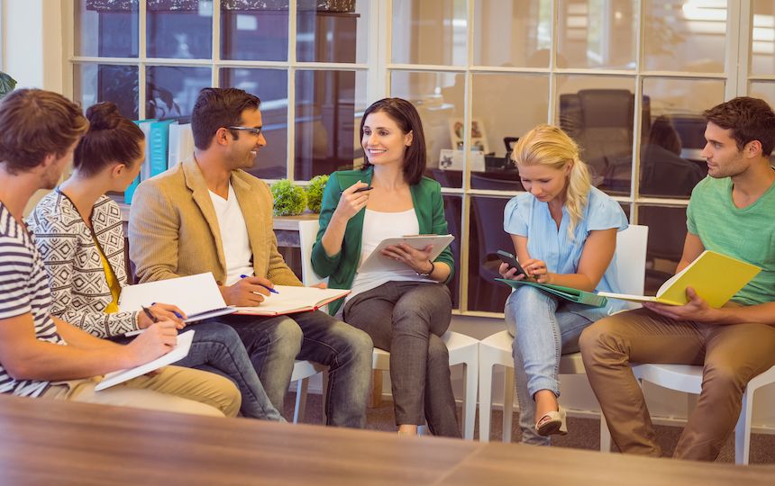 Six men and women sit in a semi-circle in an office setting, holding notepads and discussing ideas.