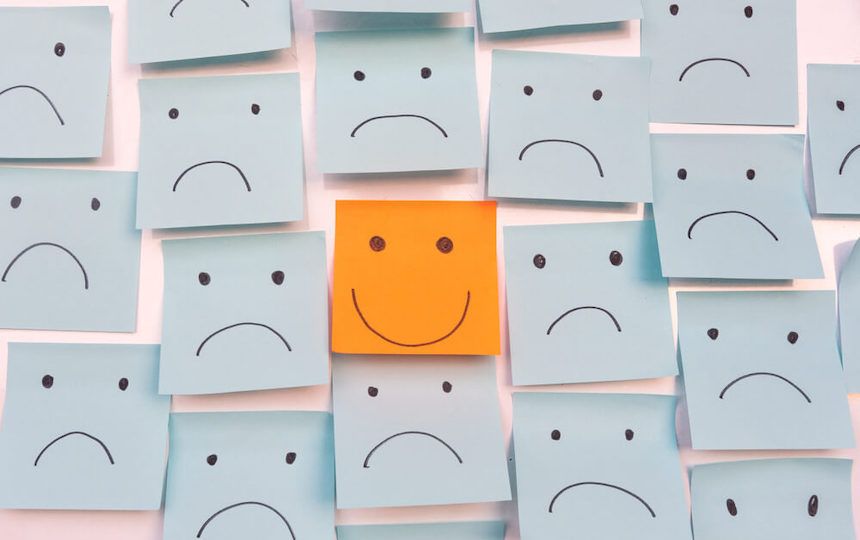 Numerous blue sticky notes with a sad face placed surround one orange note with a smile.