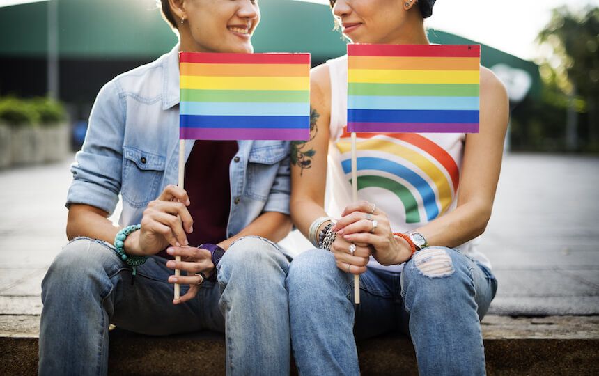 Two women sat outside on a stone step, holding rainbow flags to represent LGBT support.