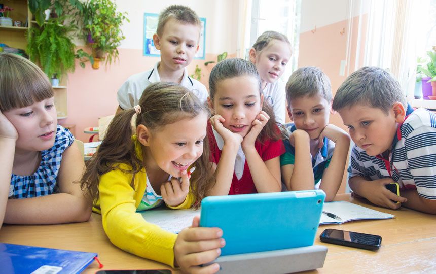 A group of young school children, some holding mobiles, playing with a tablet computer.