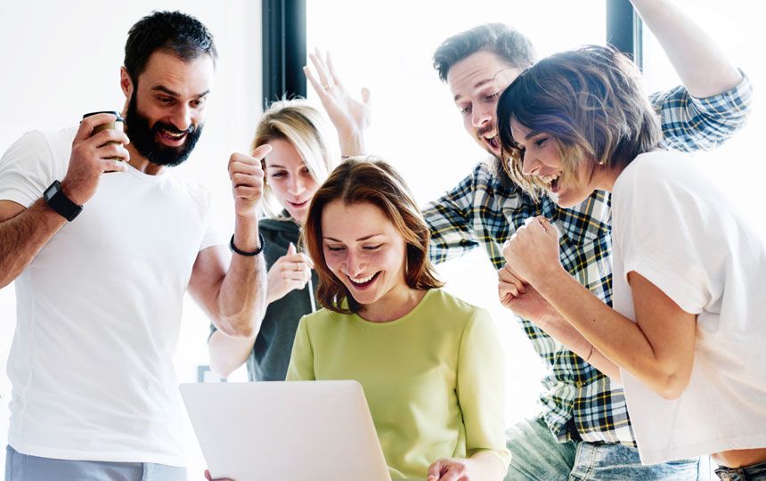 A woman smiles at her laptop whilst a group of people around her cheer happily.