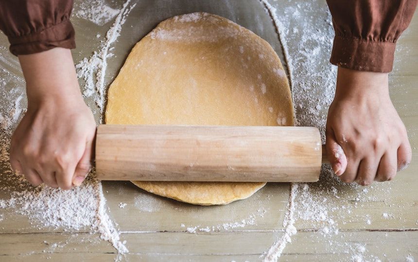 Seen from above, hands are using a rolling pin to roll out dough on a floured surface.