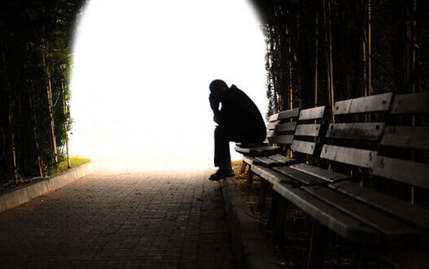 A man sits on a bench with his head in his hands against a bright natural light.