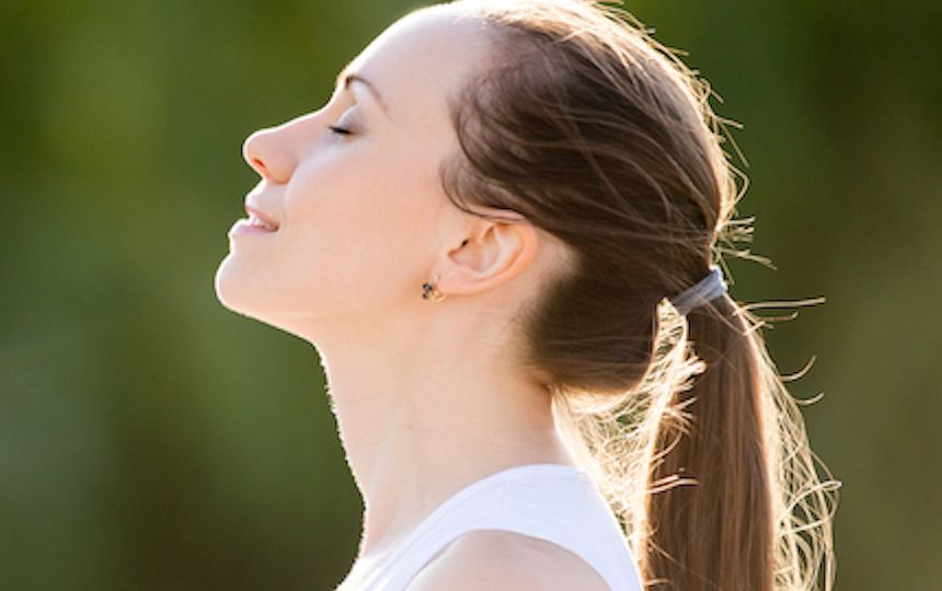 A young woman stands with her eyes closed, smiling and looking up at the sun, looking peaceful.
