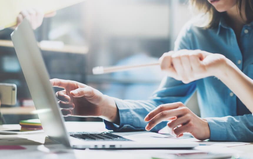 Two people discuss work on a laptop whilst one points to the screen using a pencil.