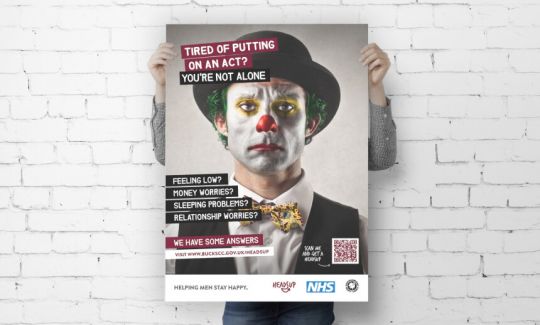 A man holds up a campaign poster of a sad-looking clown in front of a white brick wall.