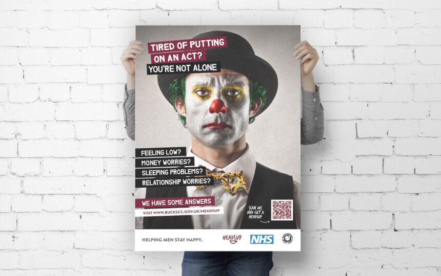 A man holds up a campaign poster of a sad-looking clown in front of a white brick wall.