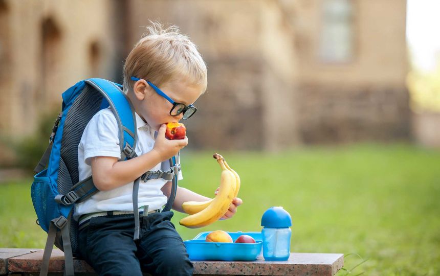 A young boy with glasses and a blue backpack is sat holding bananas and eating a nectarine.