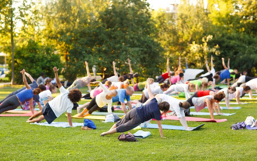 A crowd of people are using yoga mats in a park setting, performing a yoga stretch.