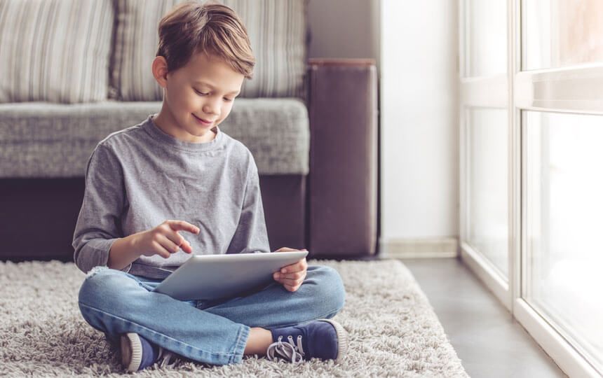 A young boy is sat on a rug at home smiling down at an iPad in his hands.