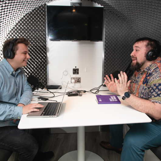 Two people sat in the pod doing a podcast wearing headphones.