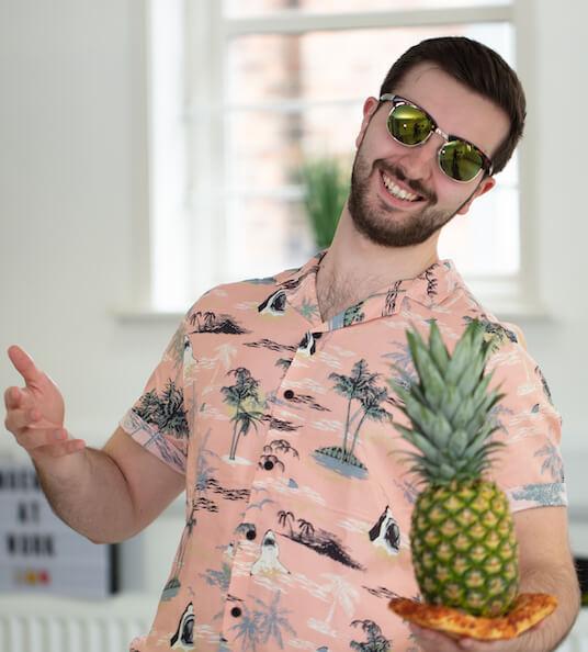 Jake, wearing sunglasses and a Havana shirt, holds a slice of pizza with a pineapple on top.