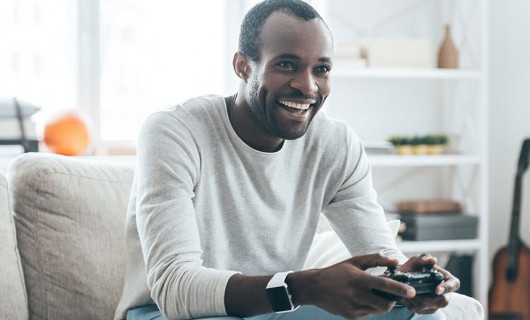 A man is sat on a sofa, holding a console remote controller and smiling.