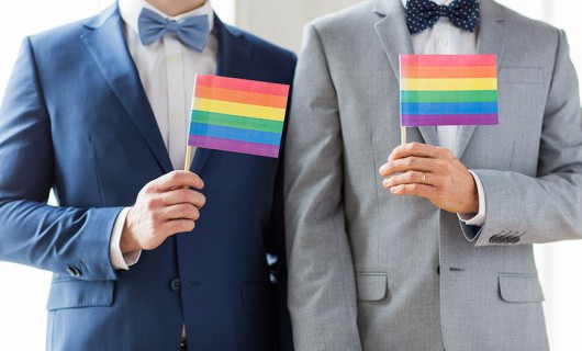 Two men dressed in smart suits holding multicoloured or rainbow coloured flags.