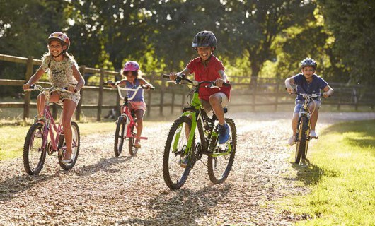 Four children are smiling and laughing whilst riding their bikes on a gravelly path.