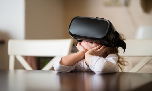 A young girl is sat at a white table, using a virtual reality headset.