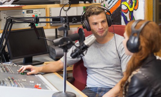 A man wearing large headphones is sat in a radio studio is talking to a lady wearing headphones.