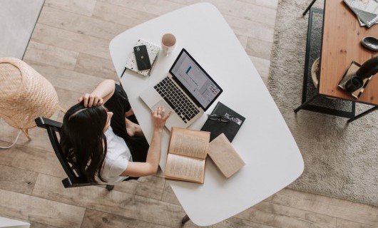 A birdseye view of a woman working on their laptop at a table with an open book.